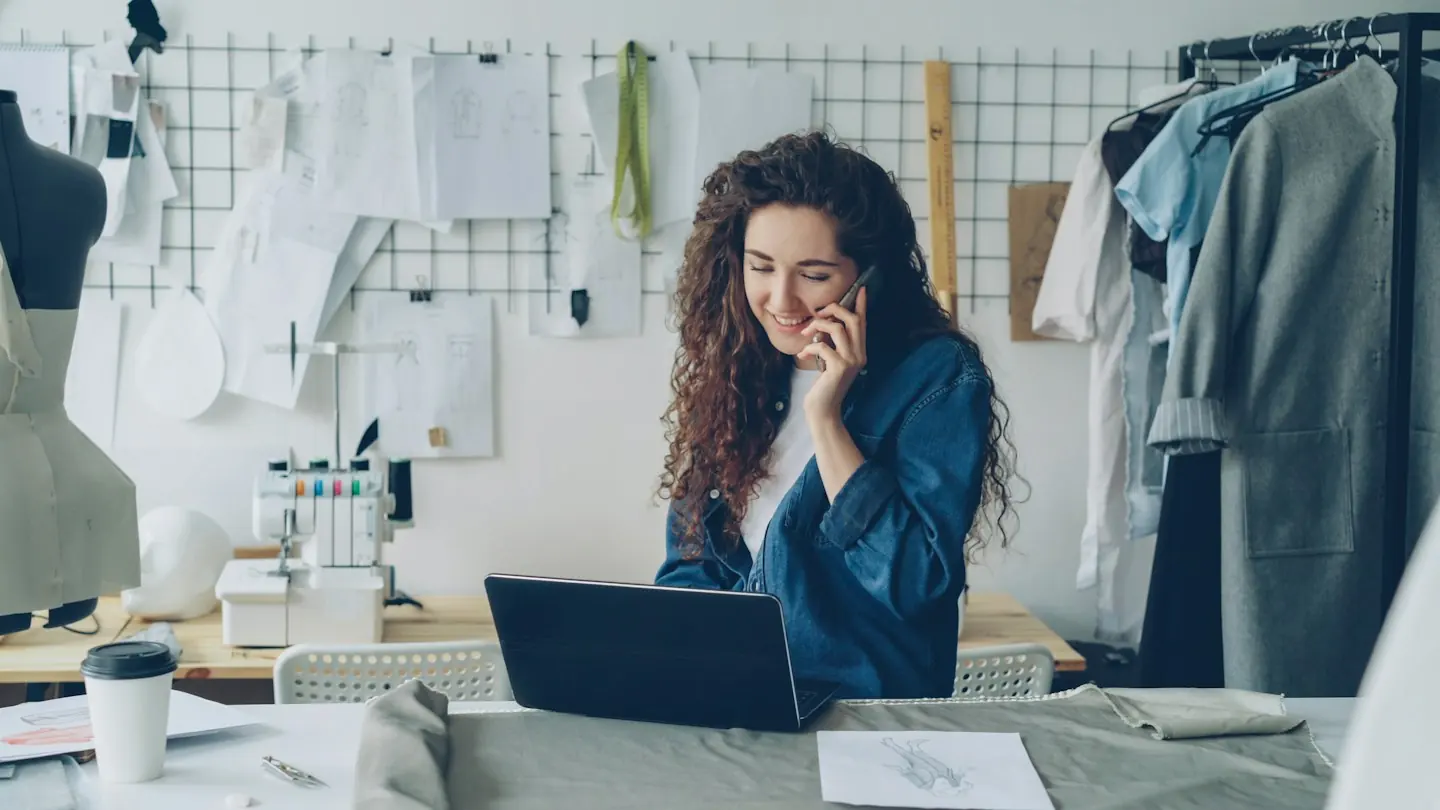 Une créatrice au téléphone devant son ordinateur portable, dans son atelier de vêtements