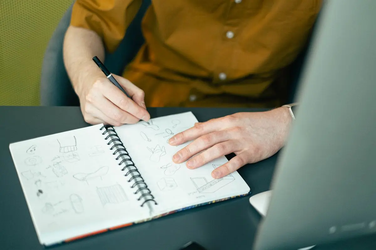 Une personne dessine des idées dans un carnet à spirale, devant un écran d’ordinateur