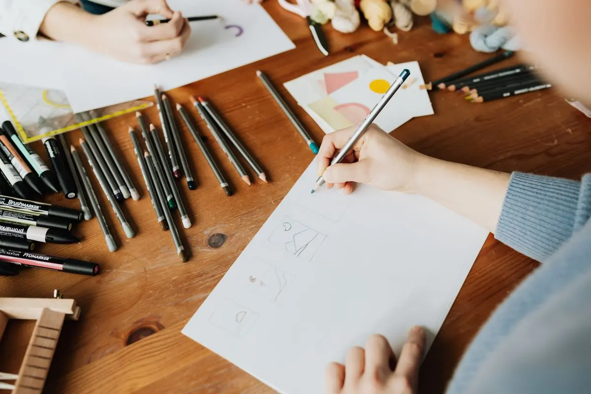 Vue du dessus : une main dessine un croquis au crayon sur une feuille, entourée de feutres et de crayons sur une table en bois