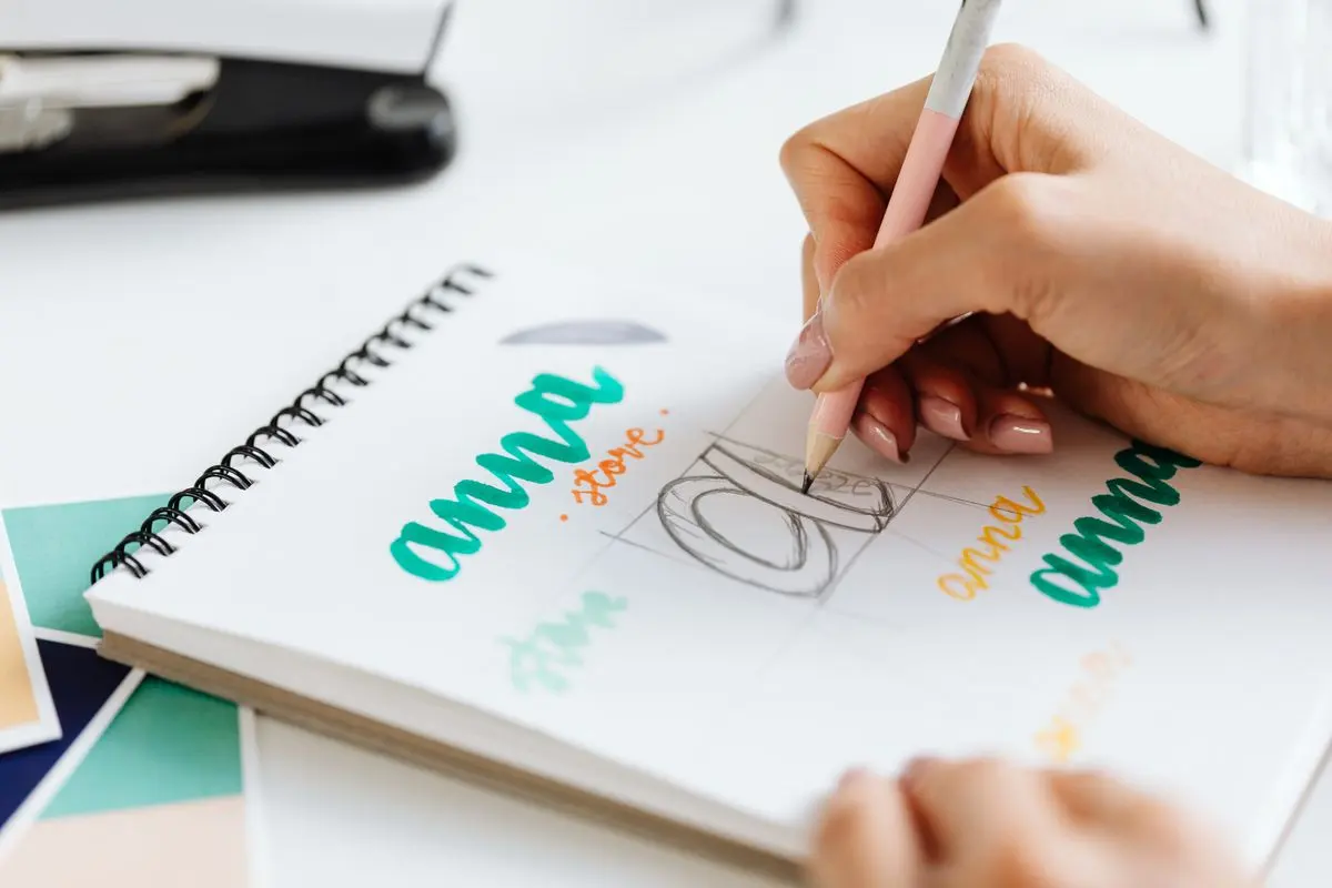 Une main trace une lettre au crayon dans un carnet de recherche de lettrage, avec des mots colorés autour