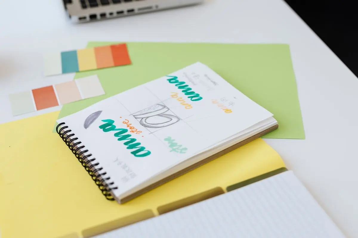 Carnet à spirale ouvert avec des recherches de lettres colorées, posé à côté de nuanciers de couleurs sur un bureau